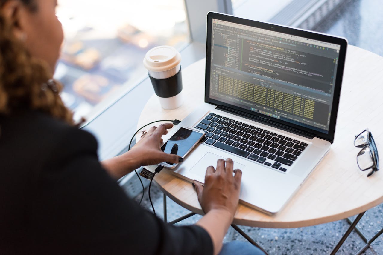 GYG CAPACITACIÓN IMPLEMENTA CAMPUS VIRTUAL Black woman programming on a laptop with coffee, smartphone, and glasses on a desk in an office.
