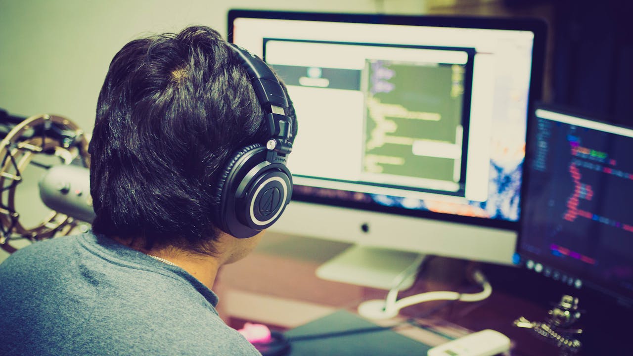 GYG CAPACITACIÓN DESARROLLA NUEVOS CURSOS PARA EL PERÍODO 2026 A programmer with headphones focuses on coding at a computer setup with dual monitors.